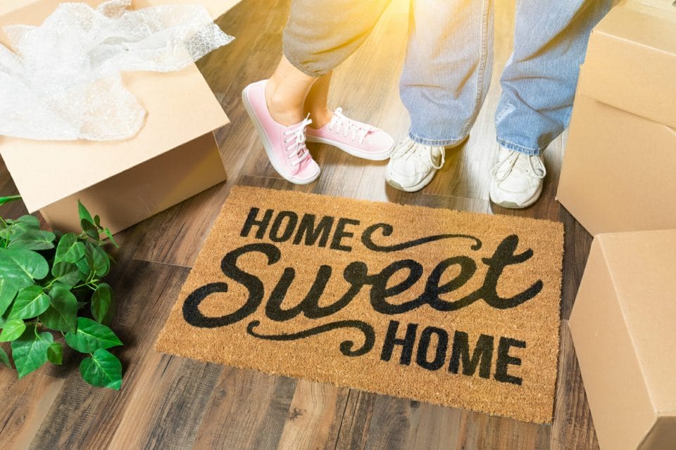 kids standing by a "Home Sweet Home" doormat in a new house